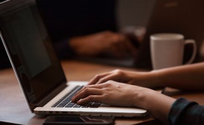 A photo of hands typing on the keyboard of a laptop, with a coffee cup in the background.